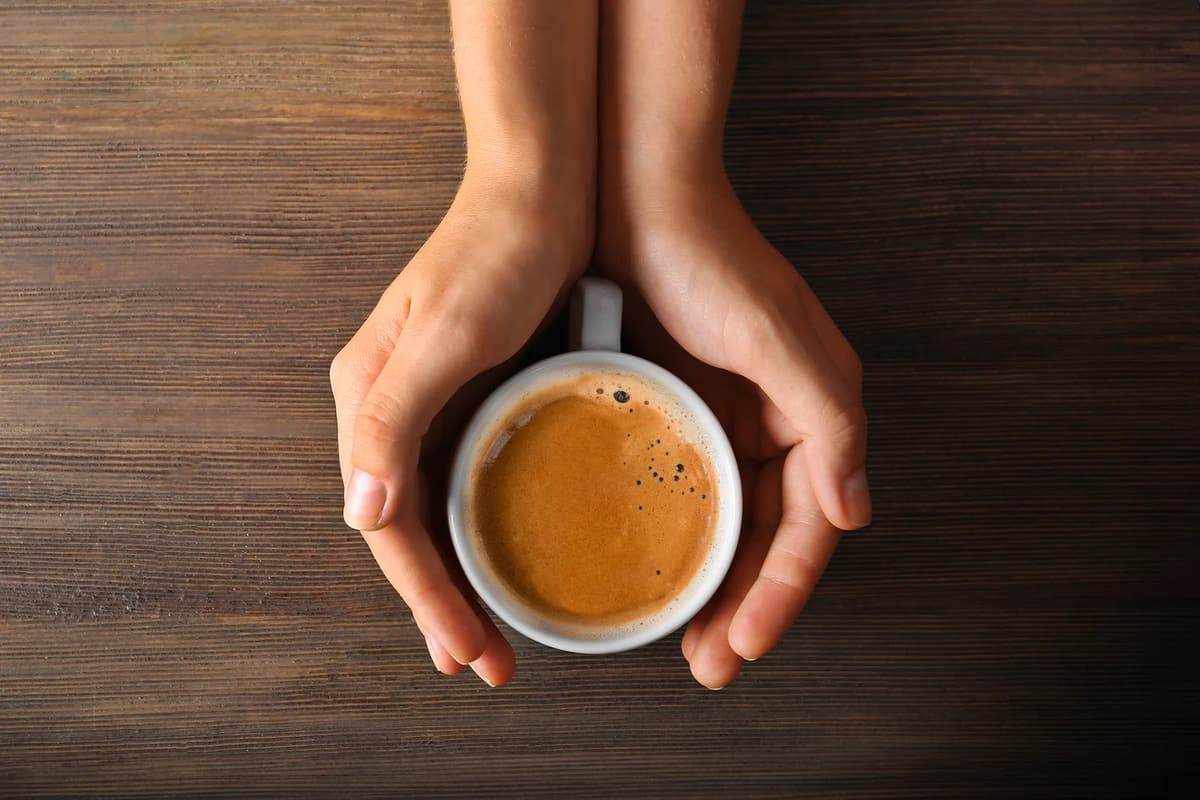 Due mani stringono una tazzina di espresso appena fatto, su un piano di legno scuro